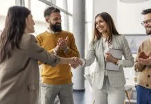 The Art of the Right Match Lies in Looking Past the Checklist: Julie Podolec’s Pursuit of Meaningful Connection Two successful female executives shaking hands for partnership, connection while standing at office while team members clapping hands at them.