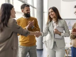 The Art of the Right Match Lies in Looking Past the Checklist: Julie Podolec’s Pursuit of Meaningful Connection Two successful female executives shaking hands for partnership, connection while standing at office while team members clapping hands at them.