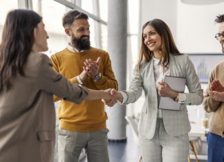Two successful female executives shaking hands for partnership, connection while standing at office while team members clapping hands at them.