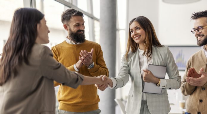 The Art of the Right Match Lies in Looking Past the Checklist: Julie Podolec’s Pursuit of Meaningful Connection Two successful female executives shaking hands for partnership, connection while standing at office while team members clapping hands at them.