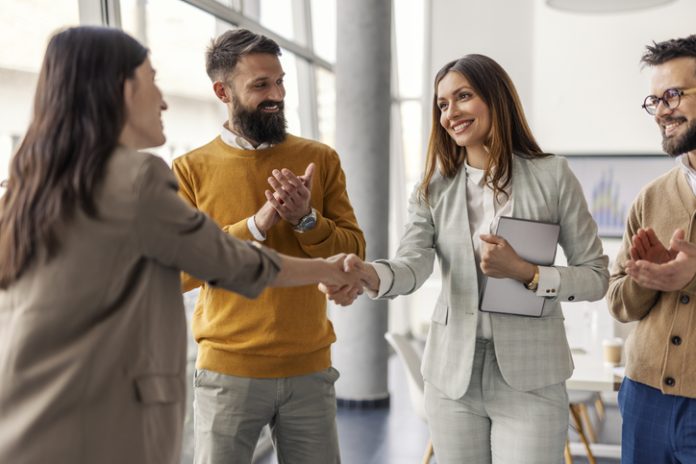 Two successful businesswomen shaking hands and closing deal and partnership while businessmen applauding them. Two successful female executives shaking hands for partnership, connection while standing at office while team members clapping hands at them.