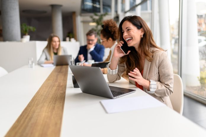 Cheerful businesswoman talking over smart phone while sitting with laptop at conference table during business meeting marketing channel through phone calls. Woman talking over smart phone while sitting with laptop