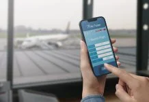 User holding a smartphone and booking cheap flights online, seasonal travel trends, airport in the background.