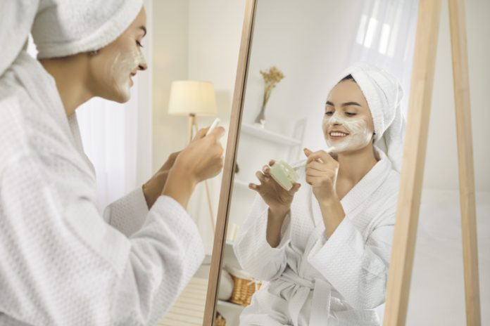 Young woman look in mirror do face mask Smiling girl after shower do morning beauty face procedures, take care of skin. Skincare and cosmetology concept.