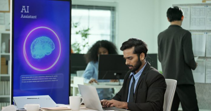 Mixed race Businessman, programmer working on a computer while looking at AI assistant interface on monitor at office. Smart workplace automation, future software engineering, machine learning concept. CEO AI strategy