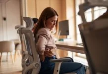 The Hidden Business Cost of Poor Ergonomics in Physically Demanding Work A woman sitting in an office chair experiencing back pain discomfort in a workplace setting. Poor Ergonomics in Physically Demanding Work