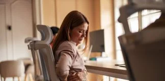 The Hidden Business Cost of Poor Ergonomics in Physically Demanding Work A woman sitting in an office chair experiencing back pain discomfort in a workplace setting. Poor Ergonomics in Physically Demanding Work