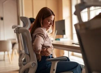 A woman sitting in an office chair experiencing back pain discomfort in a workplace setting. Poor Ergonomics in Physically Demanding Work