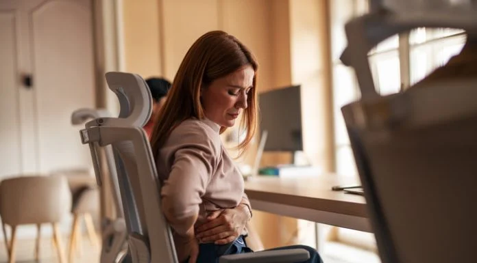A woman sitting in an office chair experiencing back pain discomfort in a workplace setting. Poor Ergonomics in Physically Demanding Work