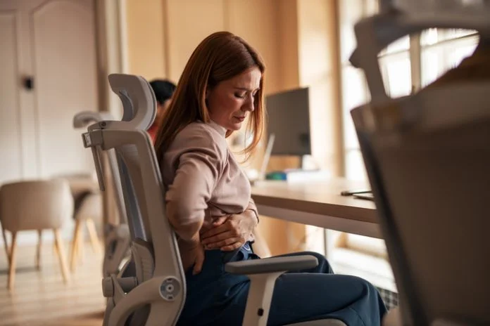 Office Worker Experiencing Back Pain While Working at Desk A woman sitting in an office chair experiencing back pain discomfort in a workplace setting. Poor Ergonomics in Physically Demanding Work