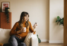 Woman relaxing on a sofa, drinking from a mug and using her phone during Free Time
