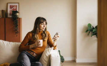 Woman relaxing on a sofa, drinking from a mug and using her phone during Free Time