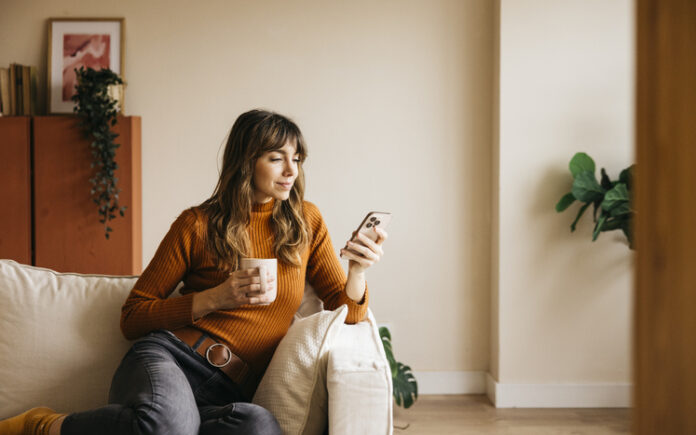 Woman relaxing on a sofa, drinking from a mug and using her phone during Free Time