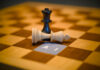 Black queen chess piece standing over a fallen white king next to a paper label reading AI, artificial intelligence dominance concept.
