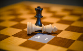 Black queen chess piece standing over a fallen white king next to a paper label reading AI, artificial intelligence dominance concept.